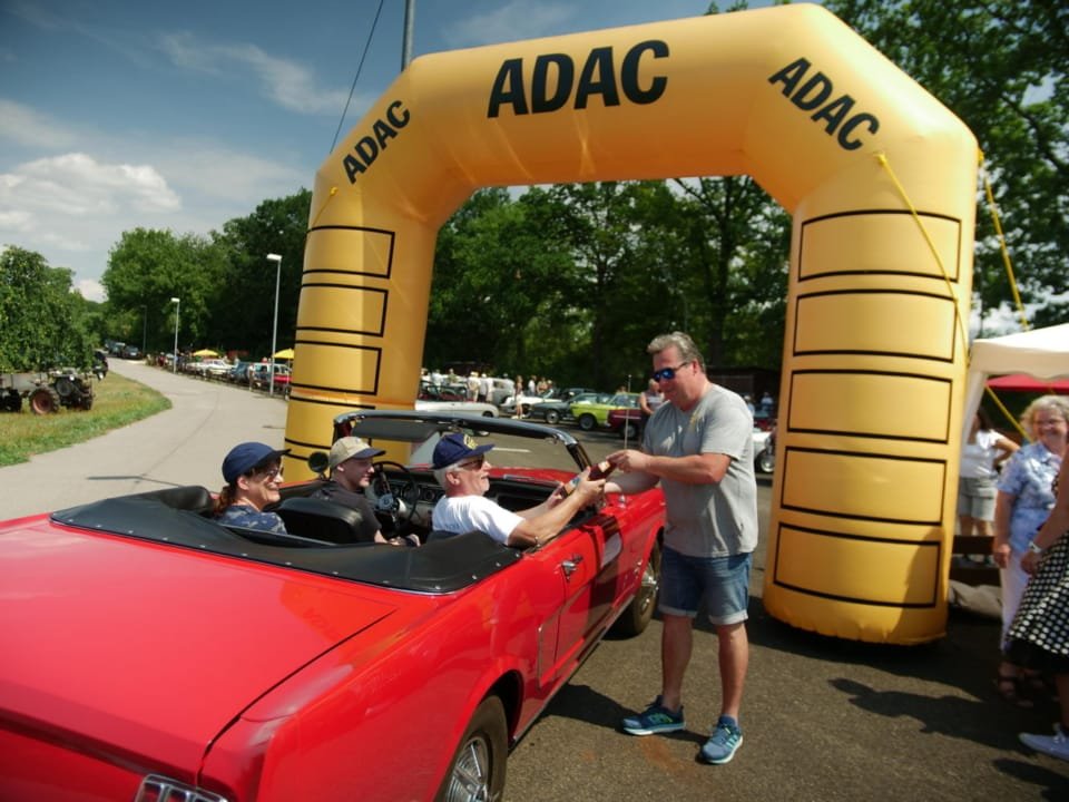 Unser Vorstand begrüßt Gäste in einem roten Muscle Car zu unserem Oldtimertreffen unter dem ADAC-Torbogen.